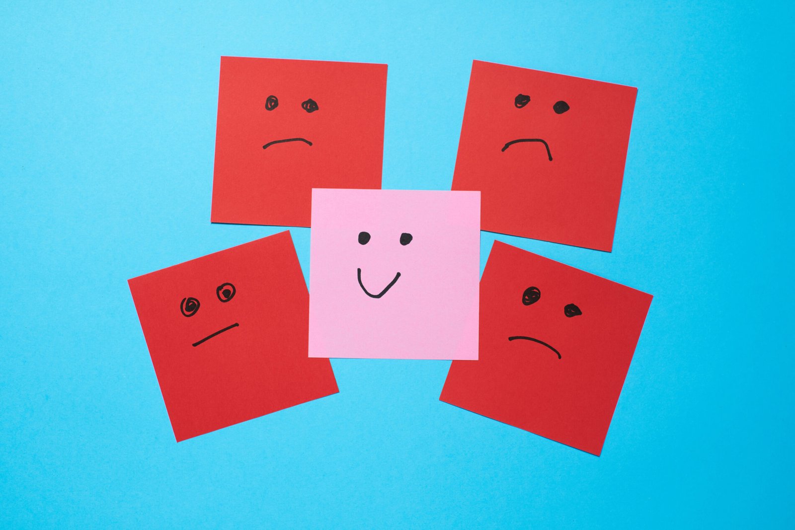 Five red and one pink paper posters are arranged on a blue background. The red posters show sad faces, while the pink poster shows a happy expression, creating a contrast.