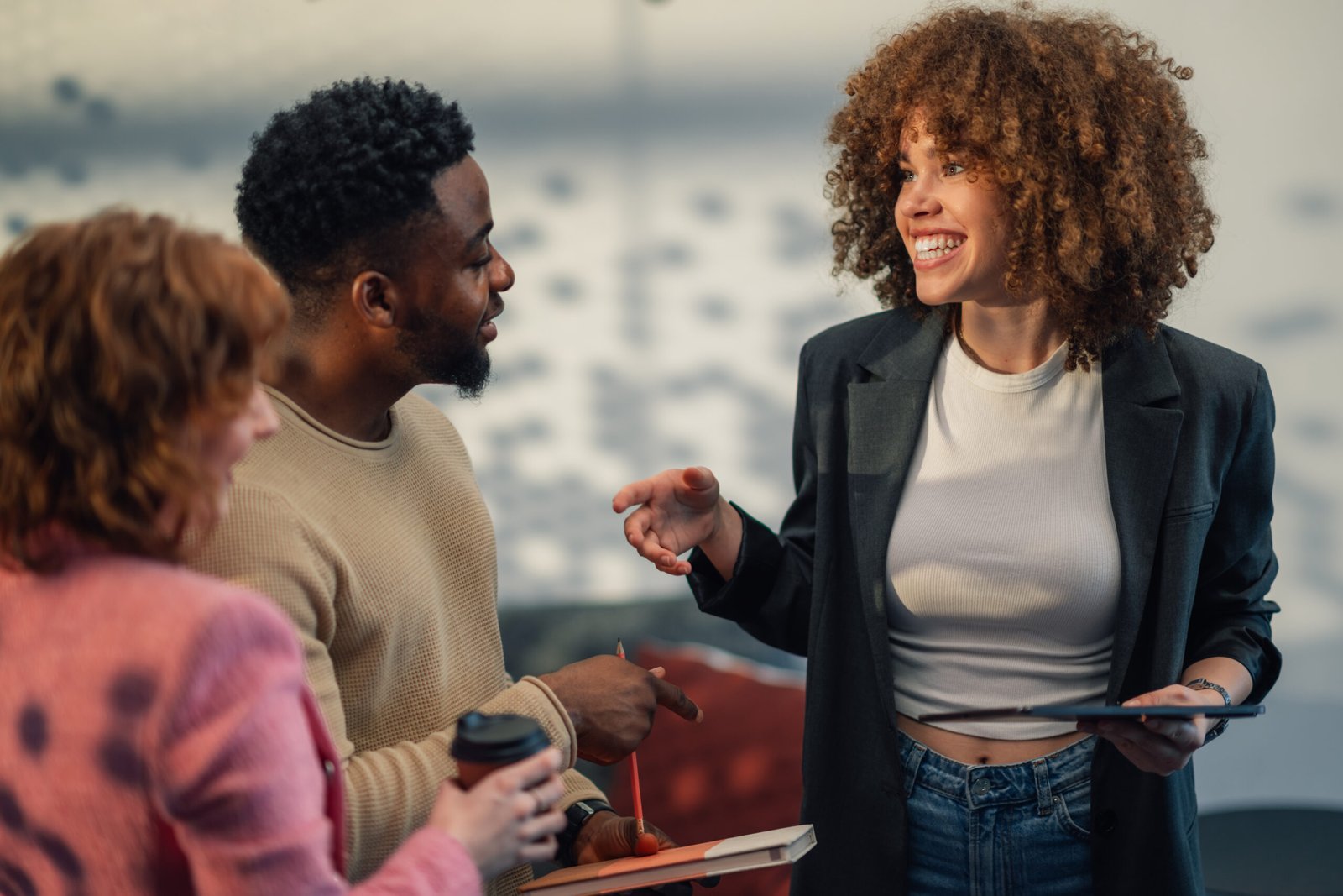 Diverse cheerful innovative business staff standing at modern office with tablet in hands and discussing business strategy at modern office. Manager with curly hair explaining project to her team.